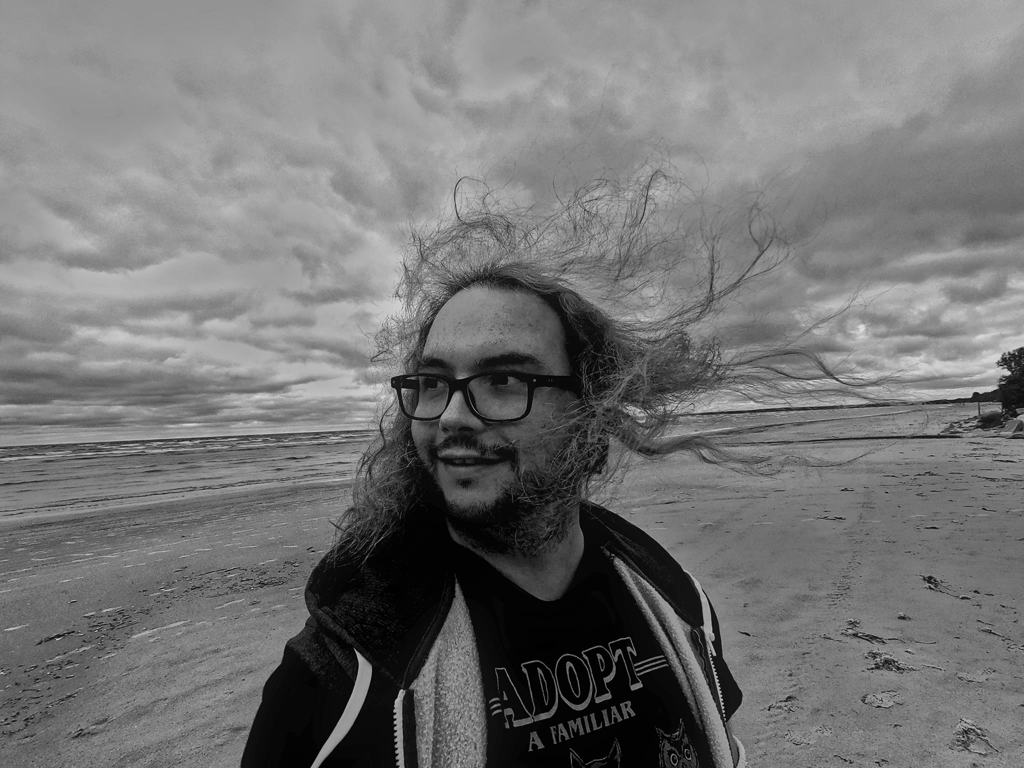 Photo is black and white, a man with long hair stands on a beach with choppy waters and a dramatic sky